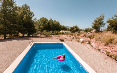 Yazın havuz manzarası. Pembe donutla süzülen mayo ve şapkalı genç kız..
