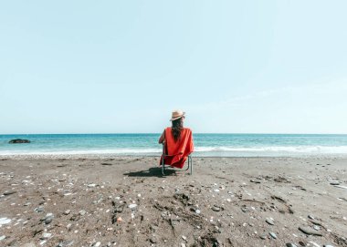 Şapkalı kız plajın kıyısında turuncu havluyla şezlongda oturuyor.