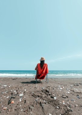 Şapkalı kız plajın kıyısında turuncu havluyla şezlongda oturuyor.