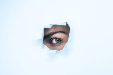 Female eye peering out of a hole in a blue paper background.