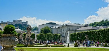 Salzburg, Avusturya - 26 Mayıs 2019: Mirabell Sarayı bahçelerindeki merkez çeşme güneşli bir sabahta ziyaretçiler tarafından keyifle dinlenir; Festung Hohensalzburg şehir simgesi yukarıdaki yüksek tüneğinden aşağıya bakar.