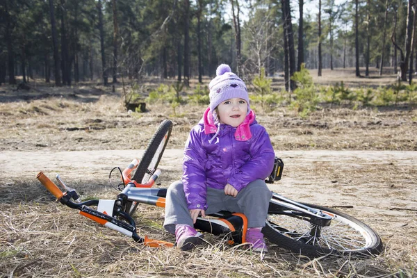 Feliz bebê engraçado menina sentada com sua bicicleta em um pinheiro — Fotografia de Stock