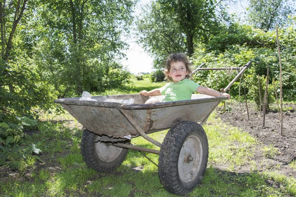 Primavera no jardim em um carrinho de mão senta-se uma menina encaracolado . — Fotografia de Stock