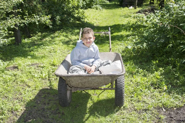 Primavera no jardim em um carrinho de mão senta-se um menino encaracolado . — Fotografia de Stock