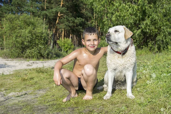 In summer, a small wet boy with a dog sits on the shore. — Stock Photo, Image