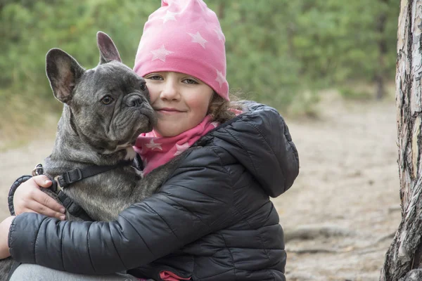 In het herfstbos houdt een klein meisje een Franse bulldog in zich. — Stockfoto
