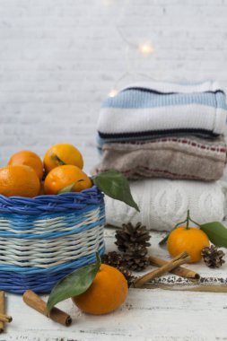 Mandalinalı Noel sepeti, tarçın çubukları ve koniler. Tahta beyaz bir arka planda. Örgü kazaklar üst üste dizilir ve çelenkler güzelce yanar..