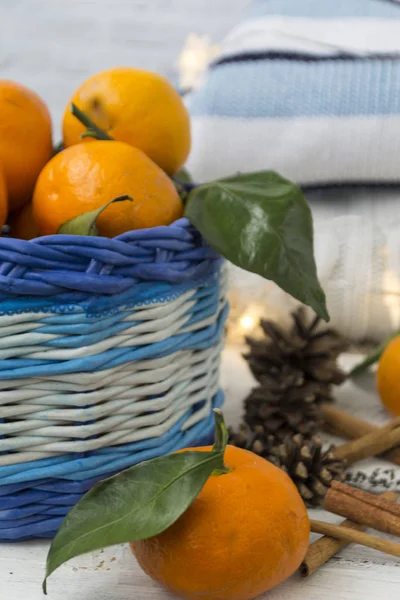 Christmas basket with tangerines, cinnamon sticks and cones. On a wooden white background. Knitted jumpers are stacked on top of each other, and a garland beautifully burns.