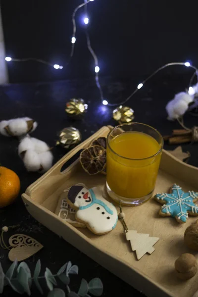 Christmas cookies in the shape of a house, a snowman and stars lie in the gap with fresh orange juice in a glass of farm-style drink. Around nuts, cinnamon, decor. Black background. — Stock Photo, Image