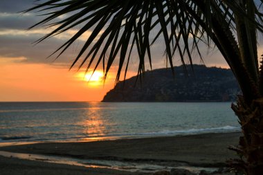 Akdeniz 'in üzerinde altın gün batımı. Batan güneş bulutların arasından parlıyor ve okyanusun üzerinde altın bir yol görünüyor. Ufuktaki siluetli dağlar. Plaj tatil yeri