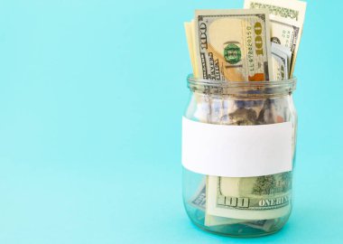 Mavi arka planda, bir litrelik kavanozdaki para. Kıyıda yazı için beyaz bir çıkartma var. Amerikan doları kumbarada. İş konsepti, finans. Metin için bir yer var.