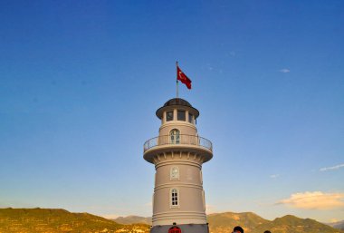 Mavi bir gökyüzüne karşı beyaz bir deniz feneri duruyor. Deniz fenerinin üzerinde kırmızı bir Türk bayrağı dalgalanıyor. Deniz fenerinin arkasında dağların ve evlerin ufuk çizgisini görebilirsiniz Deniz feneri, Türkiye, Alanya.