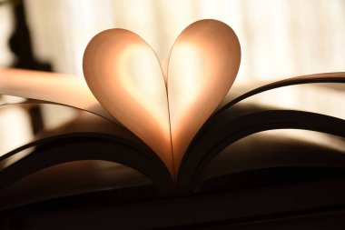 Açık sayfalı, soyut bir kitap resmi bir kalp şeklini katlamış, kitap başka kitapların üzerine istiflenmiş.