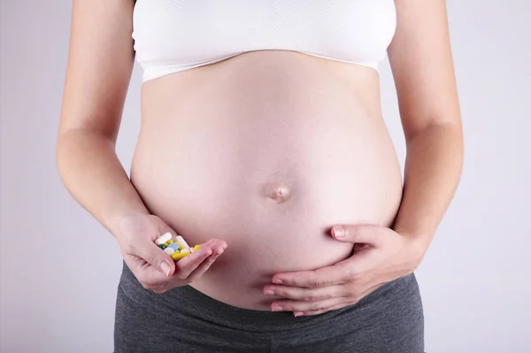 Pancia di donna incinta e pillole vitaminiche in mano su grigio — Foto Stock