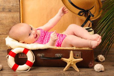 Küçük kız kahverengi arka planda plaj aksesuarlarıyla bir bavulun içinde yatıyor. Üst görünüm