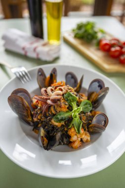 Kabuklu deniz ürünleri mürekkep balığı mürekkebi makarnası. İçinde midye karides ve ahtapot var. Arka planda yeşil ahşap bir masa üzerinde domates yağı ve sirke.