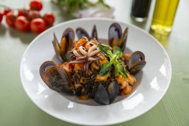 Kabuklu deniz ürünleri mürekkep balığı mürekkebi makarnası. İçinde midye karides ve ahtapot var. Arka planda yeşil ahşap bir masa üzerinde domates yağı ve sirke.