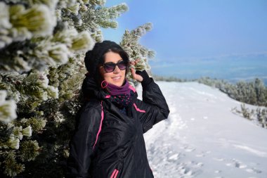 Güneş gözlüklü genç bir kadın kışın köknar ağaçlarının yanında duruyor.