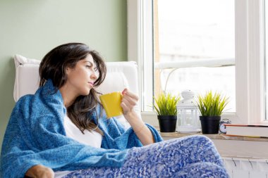 Güzel genç bir kadın, elinde bir fincan kahveyle pencereden dışarı bakıyor. 