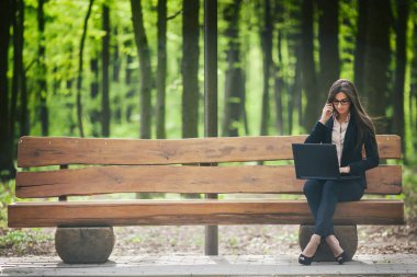 Bir dizüstü bilgisayar ile bir bankta oturmuş ve telefonda konuşurken iş kadını