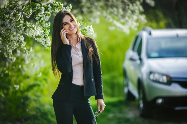 Piękna dziewczyna w pobliżu kwitnąca drzewo rozmawia przez telefon na tle samochodu — Zdjęcie stockowe