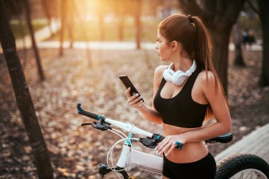 Spor kız kulaklık ile telefon ile standları ve bir bisiklet yakın mesafe içine günbatımında parkta bakıyor