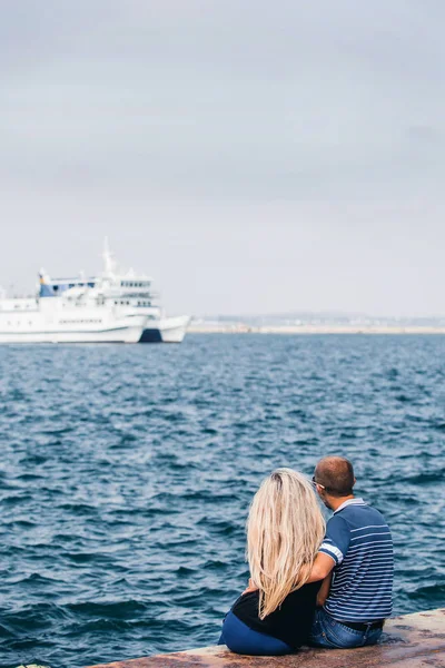 couple is sitting on the coast and looking at the ship in the sea