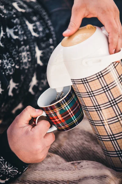 a man in a sweater with a deer pattern is holding a thermos and pouring hot tea into a cup in the winter
