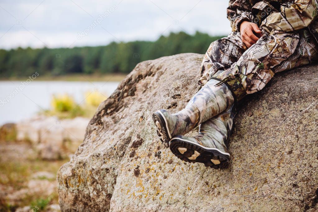 a man in rubber boots and in military uniform sits on a rock near the river in summer