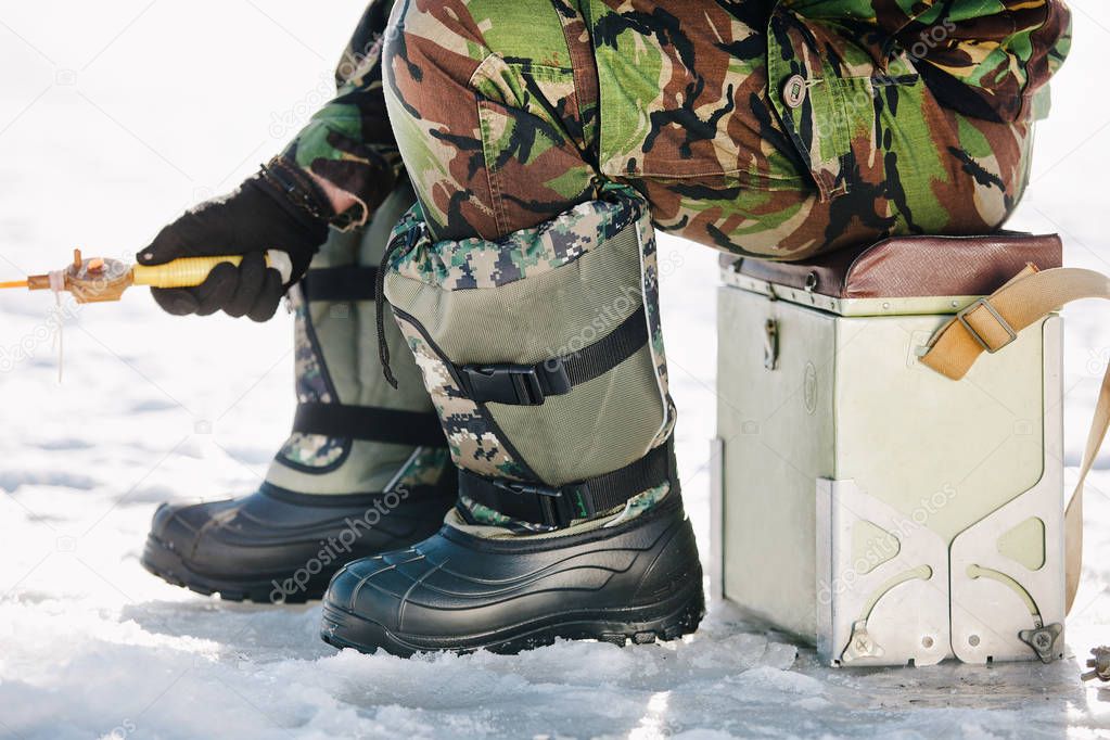a man in uniform with a fishing rod sits in felt boots on a chair on the ice of the river in the winter