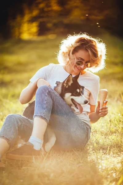 blonde with ice cream in jeans and t-shirt is sitting on the grass and holding husky in the summer in the park