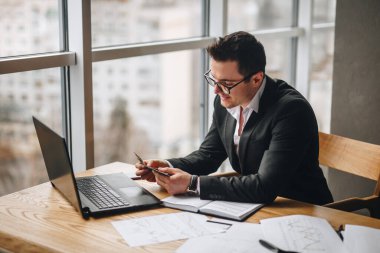 Siyah ceketli iş adamı ofiste oturuyor ve elinde bir şey tutuyor.