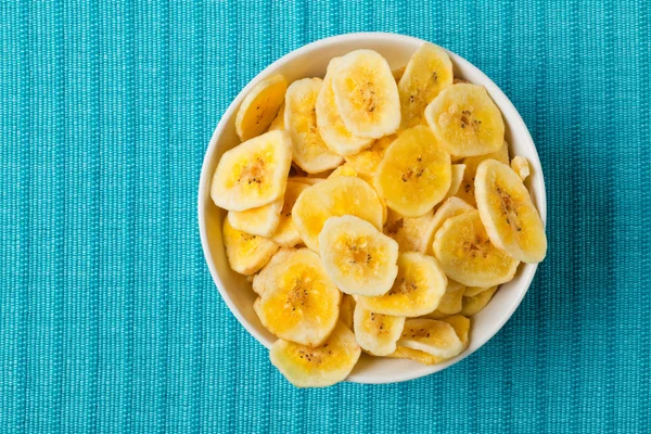 Heap of dried banana chips snack in white bowl on blue turquoise — Stock Photo, Image