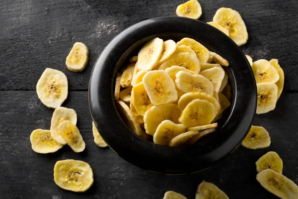 Heap of dried banana chips snack in black bowl on rustic table — Stock Photo, Image