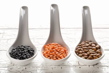 Farklı çeşitteki mercimekler beyaz tahta masa arka planında beyaz kaşıklarda kırmızı, kahverengi ve siyah mercimeklerle karıştırılır - seçici odak noktası