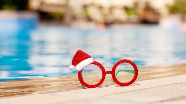 Gafas de Navidad rojas con un dibujo de Santa Claus cerca de la piscina. concepto de vacaciones de Navidad . — Foto de Stock