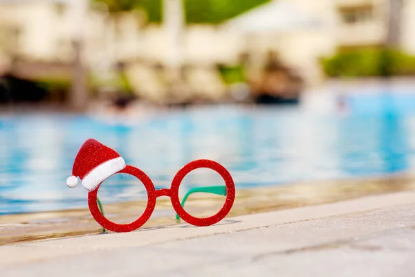 Gafas de Navidad rojas con un dibujo de Santa Claus cerca de la piscina. concepto de vacaciones de Navidad . — Foto de Stock
