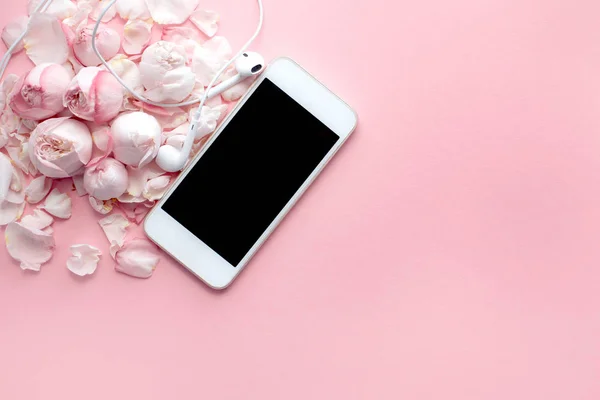 Beyaz akıllı telefon ve kulaklıklar narin güllerin ve pembe arka planda yaprakların üzerinde yatar. Üst manzara, düz yatış. Metin ve minimalizm için boşluk