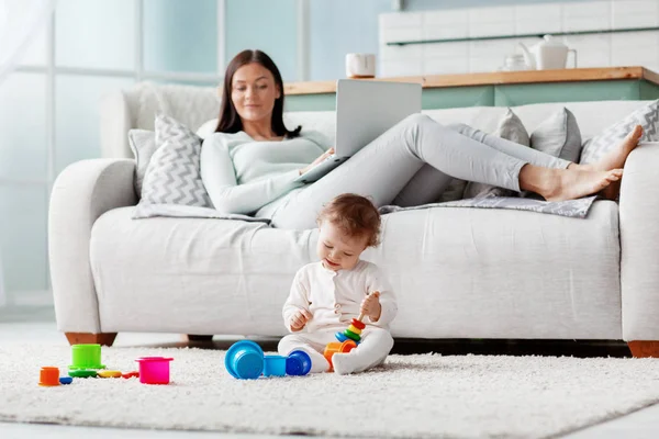 Mom works at a laptop at home while her child plays with toys on the floor. Business and family concept.