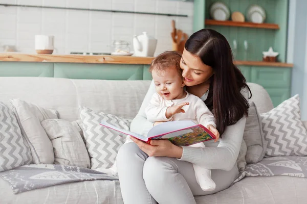 Mamma sitter på en soffa med ett barn, läser en bok och tittar på ljusa bilder — Stockfoto
