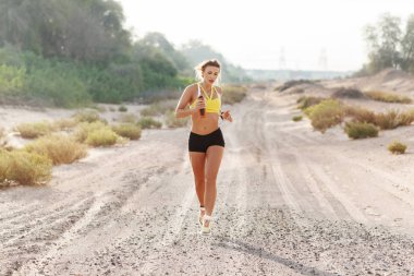 Spor giyimli genç bir kadın güneşli bir günde ağaçların arka planında kumların üzerinde koşuyor. Fitness, antrenman, yaşam tarzı