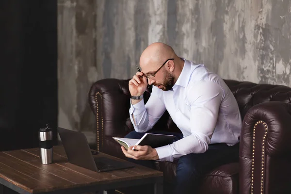 A businessman checks information in a notebook while working with a laptop in the office. A serious man with glasses writes data in a notebook. Business portrait, business, work