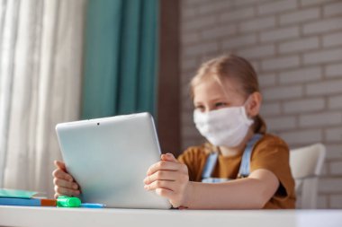 Tıbbi maskeli bir kız evde dijital tablet bilgisayarla ders çalışıyor ve ödev yapıyor. Uzaklık öğrenimi çevrimiçi eğitim