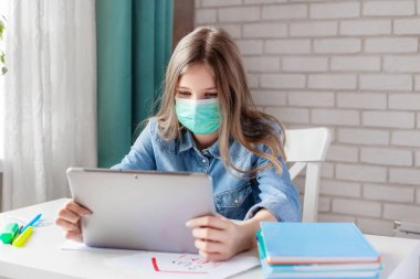 Tıbbi maskeli bir kız evde dijital tablet bilgisayarla ders çalışıyor ve ödev yapıyor. Uzaklık öğrenimi çevrimiçi eğitim