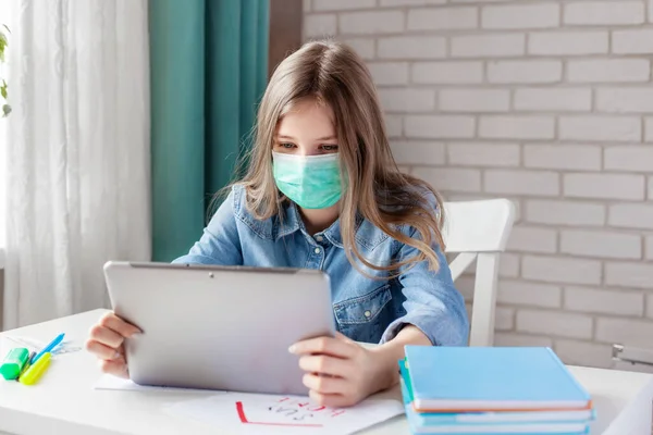 Tıbbi maskeli bir kız evde dijital tablet bilgisayarla ders çalışıyor ve ödev yapıyor. Uzaklık öğrenimi çevrimiçi eğitim