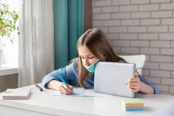 A beautiful girl in a medical mask studies at home with a digital tablet laptop and does homework, writes the answer in a notebook. Distance learning, online education