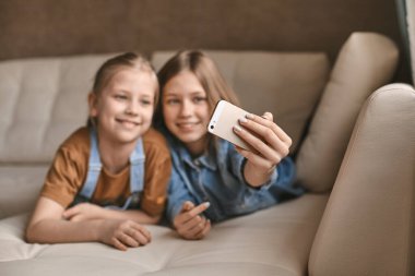 Güzel kızlar kanepede yatarken telefonlarında selfie çekerler. Kardeşler ev ödevlerine ara verip karantina sırasında fotoğraf çekerler.