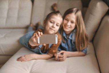 Güzel kızlar kanepede yatarken telefonlarında selfie çekerler. Kardeşler ev ödevlerine ara verip karantina sırasında fotoğraf çekerler.