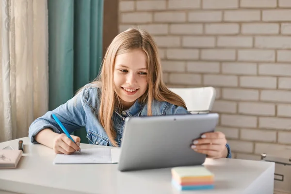 Sevimli liseli kız evde tabletle ödevini yapıyor. Çocuk öğrenmek için aletler kullanıyor. Eğitim, uzaktan eğitim, karantina sırasında evde eğitim