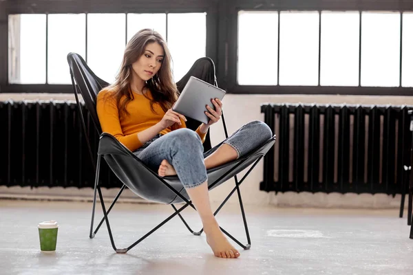 A young girl sits at home in a stylish black leather chair, holds a cup of coffee and a tablet in her hand. Work and relaxation at home